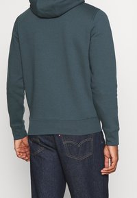 Sudadera con capucha de color verde azulado oscuro, de ajuste relajado, con capucha ajustable con cordón, puños acanalados y textura suave, combinada con jeans de mezclilla índigo.