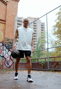 Completo sportivo da uomo con una maglietta bianca senza maniche e pantaloni corti neri. Le scarpe da ginnastica sono nere con dettagli bianchi e rosa. Pallone da basket in mano.