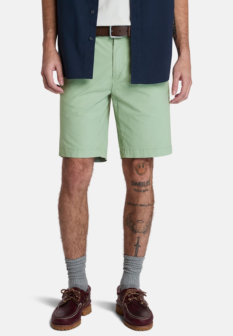 Jasnozielone szorty wykonane z bawełny, z płaskim przodem i bocznymi kieszeniami, w połączeniu z granatową koszulą na guziki i brązowym paskiem.