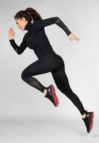 Schwarzes Sportoberteil und Leggings mit Netzeinsätzen und reflektierenden Akzenten. Schwarze und rote Laufschuhe mit strukturiertem Sohle, in einer aktiven Pose.