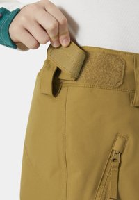 Beige broek met klittenbandsluiting en zijzak met rits; stof lijkt duurzaam, met een gladde textuur en schone stikseldetails.
