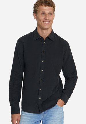 Mann lächelt, trägt ein schwarzes Hemd mit langen Ärmeln und hellblaue Jeans, eine Hand in der Tasche vor neutralem Hintergrund.