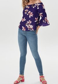 Violetti olkaimeton paita, jossa on kukkakuosi, levenevät hihansuut, yhdistettynä vaaleansinisiin skinny-farkkuihin ja vaaleanpunaisiin sandaaleihin.