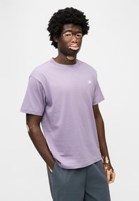 Λιλά κοντομάνικη μπλούζα από βαμβάκι, με στρογγυλή λαιμόκοψη, χαλαρή γραμμή και μικρό λογότυπο στο στήθος. Συνδυασμένη με γκρι παντελόνια.