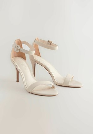Bej színű magassarkú szandál bokapánttal, sima felülettel és letisztult dizájnnal. Patentos részlet a pánton.