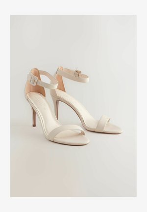 Bēšas augstpapēžu sandales ar potīšu siksnām, kurām ir gluda tekstūra un minimālisma dizains. Siksnas detaļa ar sprādzi.