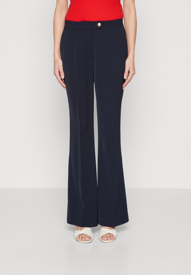 Granatowe szerokie spodnie o gładkiej fakturze, z zapięciem na guziki z przodu i paskiem, zestawione z białymi sandałami na paski.