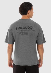 Camiseta de manga corta gris con texto negro en la parte posterior: "SMILODOX TRAINING CLUB" con frases motivacionales. Tejido suave, corte casual.