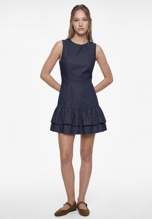 Ung kvinde står med hænderne bag ryggen iført en ærmeløs, mørkeblå lagdelt minikjole og brune flade sko.