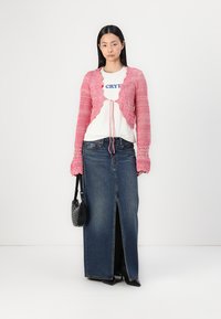 Cárdigan de punto rosa con bordes fruncidos, superpuesto a una camiseta gráfica blanca, combinado con una falda de mezclilla larga con una abertura frontal.