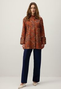 Bluză maro cu flori, cu guler și mâneci lungi, asortată cu pantaloni evazați de culoare albastru închis și pantofi cu toc în nuanțe deschise. Material neted, cu model.