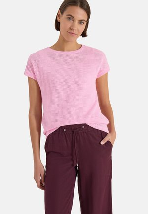 Femme portant un haut en maille rose à manches courtes et un pantalon violet foncé avec cordon de serrage, debout, une main dans la poche, sur fond blanc.