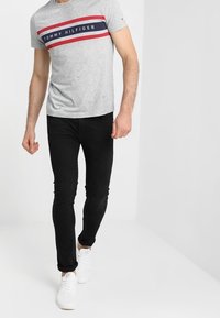 Camiseta de algodón gris con rayas rojas y marinas, texto del logo en el centro. Combinada con jeans negros ajustados y zapatillas blancas.