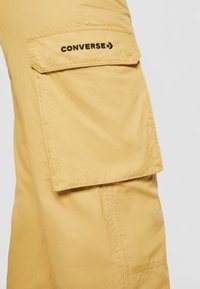 Pantalon cargo couleur beige avec une grande poche latérale à rabat brodée du logo Converse noir sur la cuisse.