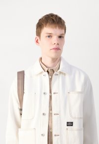 White denim jacket with visible stitching and pockets, layered over a beige plaid shirt. Model carries a brown strap backpack.