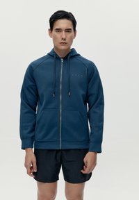 Felpa con zip color navy in tessuto morbido, con cappuccio, tasche frontali e cordino. Indossata sopra pantaloni corti neri. Dettaglio logo semplice sul petto.