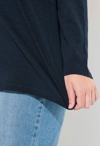 Dunkelblaues Langarmoberteil mit einem drapierten Design und einem abgerundeten Saum. Eine Hand greift den Stoff am unteren Rand, getragen mit hellblauen Jeans.