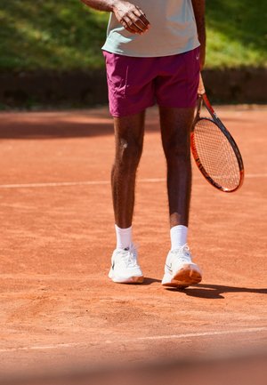 Man in witte sneakers en paarse shorts met een tennisracket staande op een klei tennisbaan.