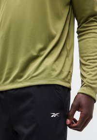 Κοντινό πλάνο ενός ατόμου που φοράει ελιά πράσινη μπλούζα μακρυμάνικη και μαύρο σορτς με λευκό λογότυπο Reebok, το χέρι ακουμπάει στο πλάι.