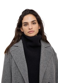 Cappotto grigio in lana con un colletto strutturato indossato sopra un dolcevita a maglia scura. Presenta una finitura testurizzata e una vestibilità rilassata.
