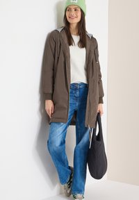 Femme portant un bonnet en tricot vert, un long manteau marron, un pull blanc, un jean bleu et des baskets, tenant un sac fourre-tout matelassé noir, appuyée contre un mur.