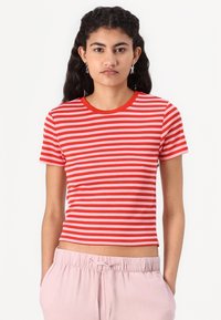 T-shirt rayé rouge et rose avec un col rond et des manches courtes, fabriqué en tissu doux. Le modèle porte un pantalon léger, rose pâle et ample.