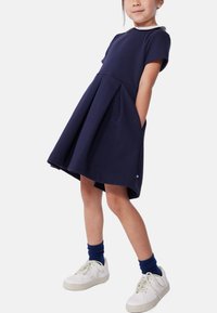 Ναυτικό φόρεμα με κοντό μανίκι, πλισέ φούστα και τσέπες. Διαθέτει λευκό γιακά και συνδυάζεται με λευκά sneakers και μπλε κάλτσες.