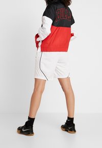 Röd och svart färgblockerad jacka med "BULLS" i stora bokstäver på ryggen, tillsammans med vita shorts och svarta sneakers.