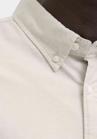 Camicia beige chiaro con bottoni, colletto strutturato, bottoni bianchi e tessuto strutturato con un motivo a trama sottile.