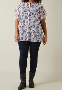 Kvinna klädd i en vit skjortblus med lila blommönster, mörka skinny jeans och svarta klackiga ankelstövlar som står mot en neutral bakgrund.