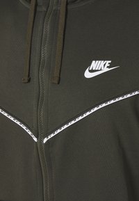 Felpa verde con zip e logo Nike bianco, tessuto testurizzato, cappuccio con cordino e una striscia bianca con branding ripetuto lungo il petto.