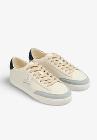 Sneakers in pelle crema con accenti azzurri, lacci beige e un tallone glitterato blu scuro. Presentano un logo con teschio e ossa incrociate.