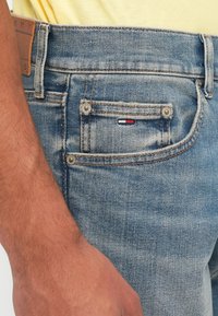 Lähivõte inimese käest sinise denimpükside tasku kõrval, millel on väike punane, valge ja sinine Tommy Hilfigeri logo ning nähtav õmblemine.