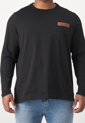 Homme portant un t-shirt noir à manches longues Calvin Klein avec un logo rouge sur la poitrine et un jean bleu clair, vue du torse.