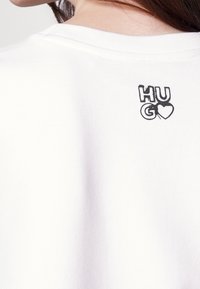 Camiseta de algodón blanca con un diseño gráfico texturizado en la parte de atrás que lleva la palabra "HUGO" y un icono de corazón en negro.
