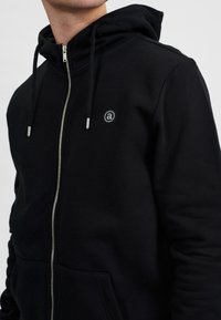 Schwarzer Kapuzenpullover mit Reißverschluss aus weichem Stoff, mit Kordelzugkapuze, silbernem Reißverschluss und kleinem Logo auf der Brust.