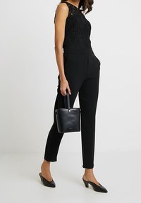 Top negro de encaje sin mangas combinado con pantalones negros ajustados, complementado con un pequeño bolso cuadrado y tacones negros de punta.