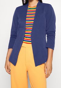 Blazer bleu marine à coupe ajustée, ouvert sur le devant, avec des manches longues. Associé à un haut à rayures multicolores et un pantalon orange.