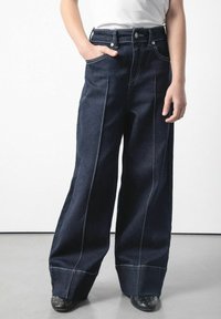 Szerokie ciemnoniebieskie dżinsy z kontrastowym przeszyciem, wysoki stan, boczne kieszenie i zwinięte nogawki. W połączeniu z czarnymi butami z ćwiekami.