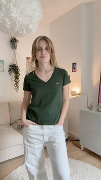 Zielony t-shirt z dekoltem w serek i małym czerwonym logo na piersi, zestawiony z luźnymi białymi spodniami, umieszczony w przestrzeni wewnętrznej w neutralnych tonacjach.