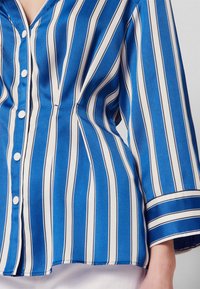 Modrá a bílá pruhovaná košile s knoflíkovým zapínáním vpředu, dlouhými rukávy a přiléhavým střihem. Materiál se zdá být hladký a lehký.