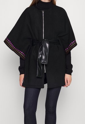 Cape noire à fermeture éclair avec bordure rose brodée sur les manches, portée par-dessus un col roulé noir, cintrée à la taille avec une ceinture en cuir noir.