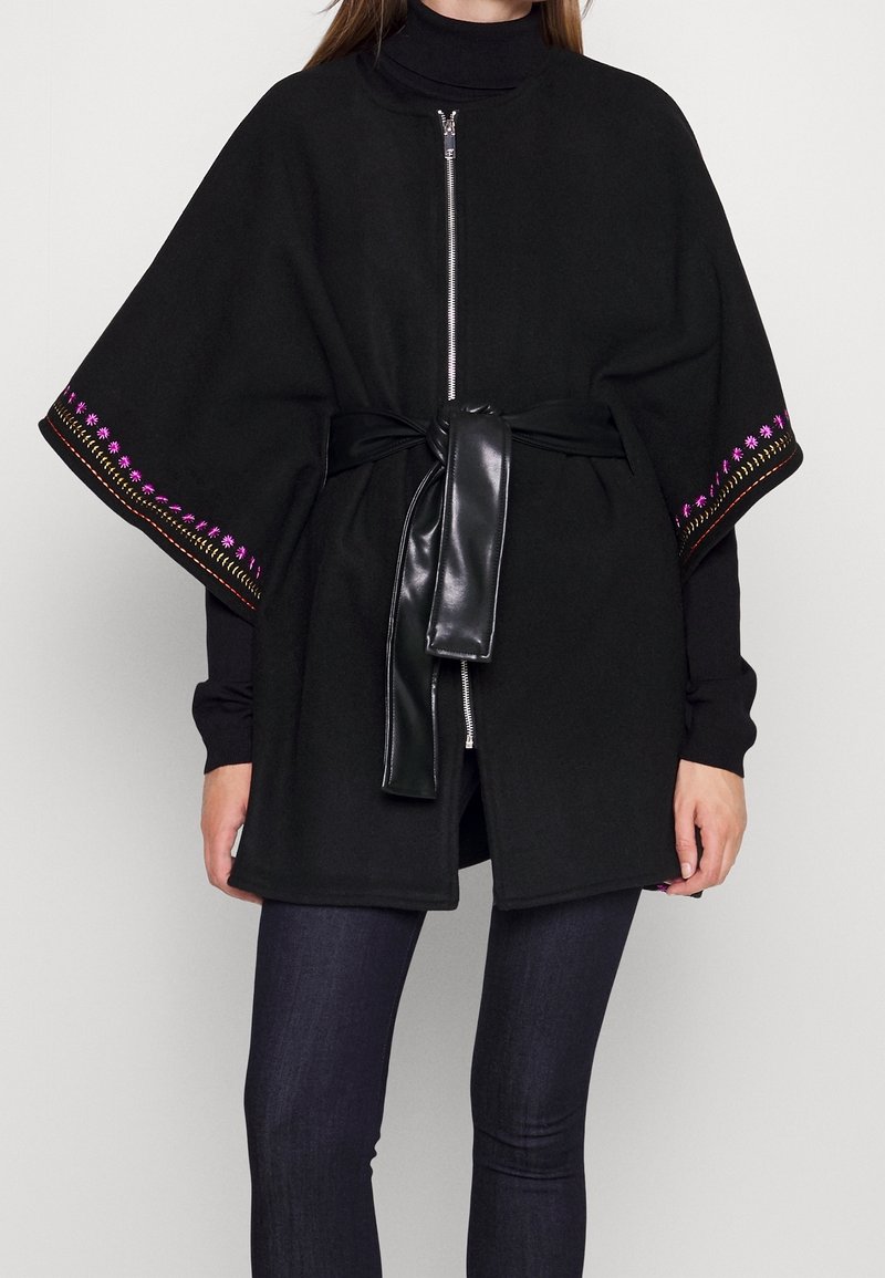 Cape noire à fermeture éclair avec bordure rose brodée sur les manches, portée par-dessus un col roulé noir, cintrée à la taille avec une ceinture en cuir noir.