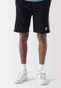 Pantalones cortos deportivos negros con rayas blancas en los lados, con una cinturilla elástica y detalle de logo en el dobladillo. Combinados con zapatillas blancas y calcetines hasta la mitad de la pantorrilla.