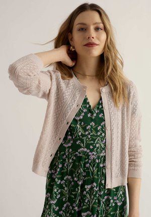 Frau mit langen blonden Haaren, die goldene Ohrringe, eine hellrosa gestrickte Strickjacke und ein grünes Blumenkleid trägt, posiert mit einer Hand hinter dem Nacken.