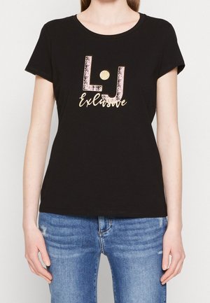 Frau trägt schwarzes T-Shirt mit kurzen Ärmeln mit pinkem und goldenem "L.J Exclusive"-Logo, kombiniert mit blauen Jeans.
