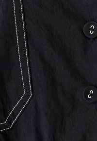 Zwarte stof met witte contrasterende stiksels, een grote knoop en een diagonale naaddetails. De textuur lijkt glad met een lichte glans.