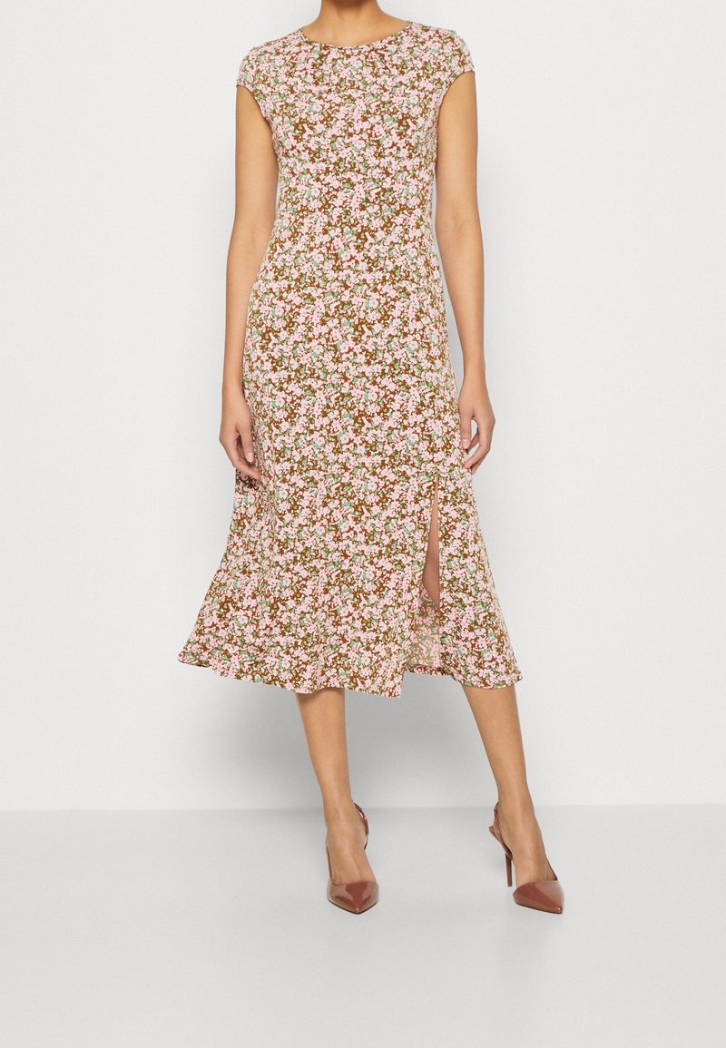 Femme portant une robe midi marron à fleurs roses, manches ballon, fente frontale et talons hauts marron pointus.