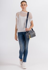 Top corto in rete bianca sopra una camicia bianca aderente, abbinato a jeans blu. Una borsa grigia con dettagli gialli è portata sulla spalla.