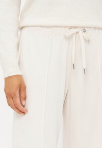 Lys beige joggebukser i et mykt, strukturert stoff, elastisk midje med snor og metallender, designet for komfort.